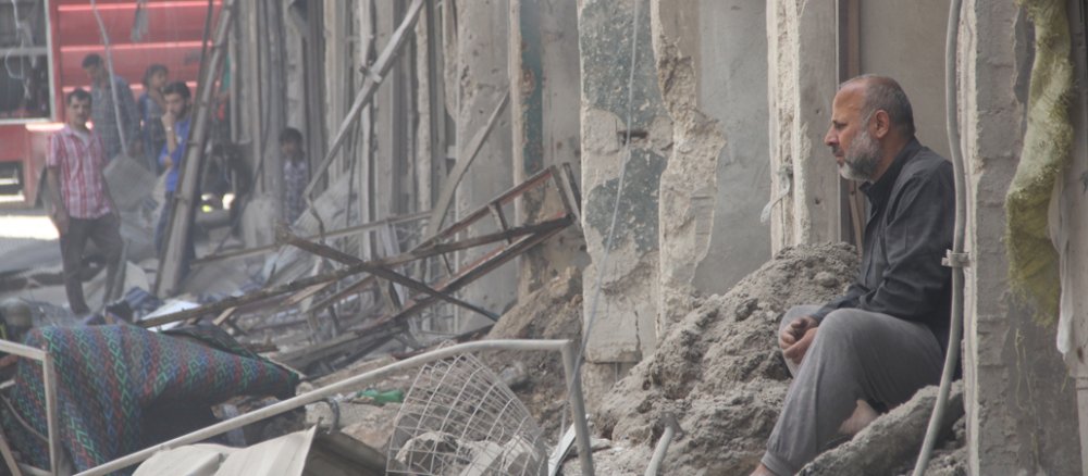 Ein älterer Mann sitzt rechts in etwas, was ein Hauseingang gewesen sein könnte. Man blickt auf eine Straße voller unaufgeräumter Trümmer in Aleppo/Syrien.