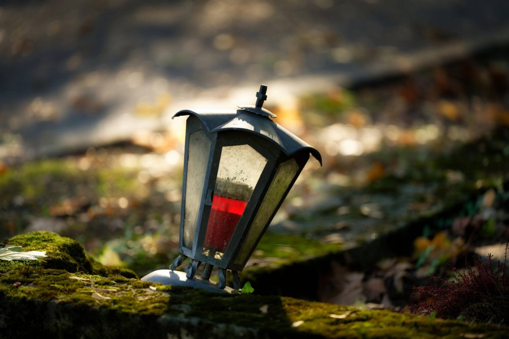 Geneigte Grablaterne mit roter Kerze im goldenen Herbstlicht auf moosigem Stein auf dem Melaten-Friedhof.