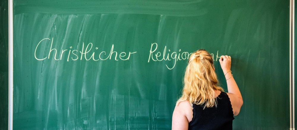 Eine Lehrerin schreibt den Schriftzug "Christlicher Religionsunterricht" mit Kreide an eine Tafel.