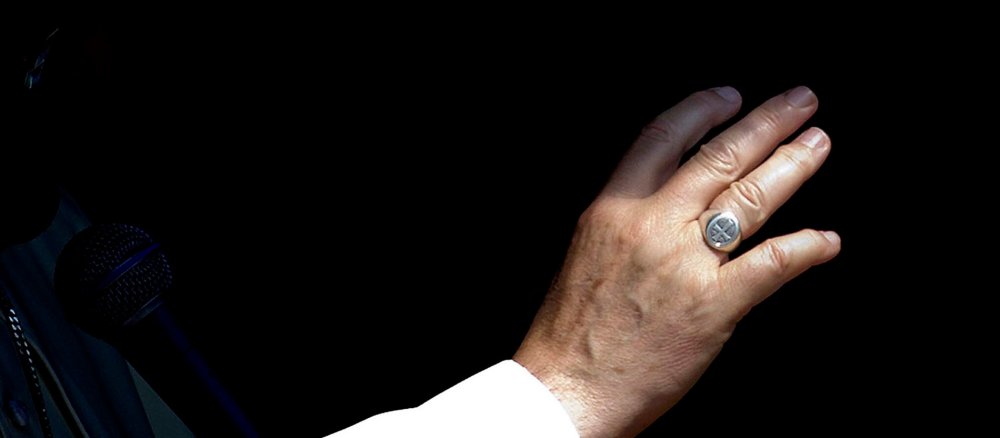 Die zum Segen ausgestreckte Hand des Papstes mit seinem Bischofsring vor schwarzem Hintergrund