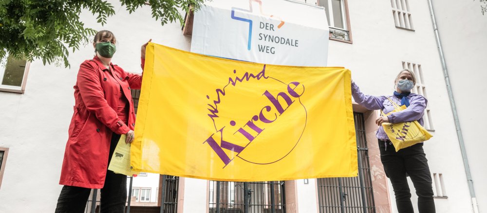 Zwei Frauen stehen mit einem Banner von "Wir sind Kirche" vor einem Gebäude, im Hintergrund ein Banner des Synodalen Wegs