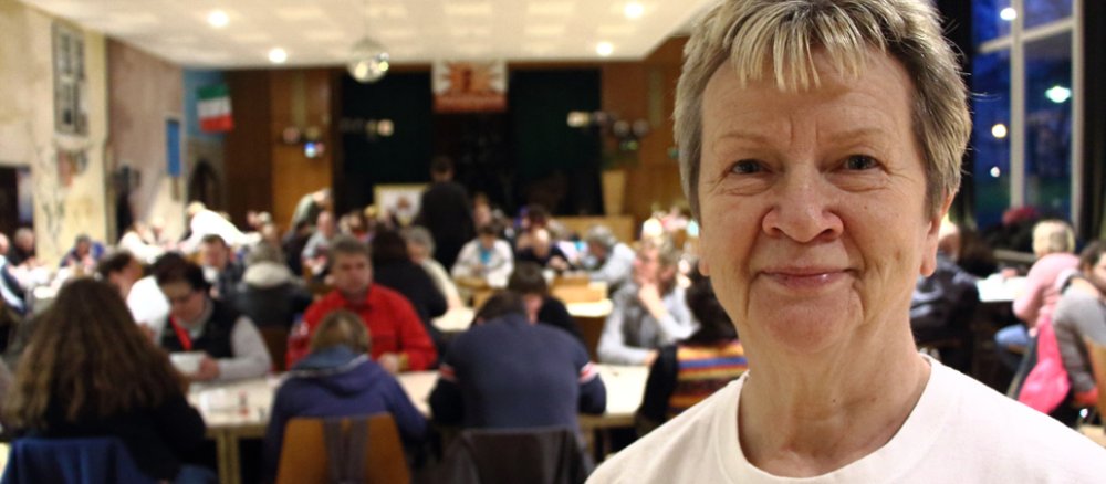 Eine Frau mittleren Alters, mit kurzen Haaren steht im großen Saal ihres Restaurants. Im Hintergrund tummeln sich unscharf die Gäste.