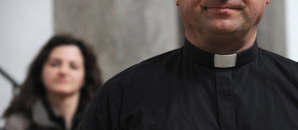 Im Vordergrund ein Priester mit Kollarkragen und im Hintergrund eine Frau.
