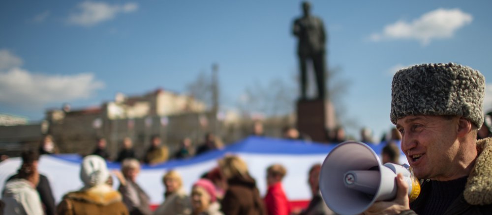 Ein Mann mit grüner Kleidung und einem Lautsprecher, dahinter unscharf Menschen mit einer russischen Flagge und eine Statue von Lenin