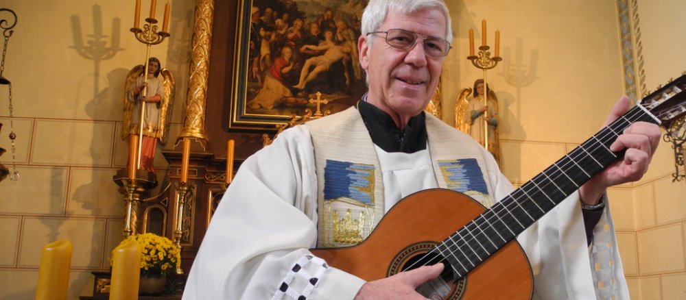 Ein Pfarrer steht mit einer Gitarre in einer Kirche.