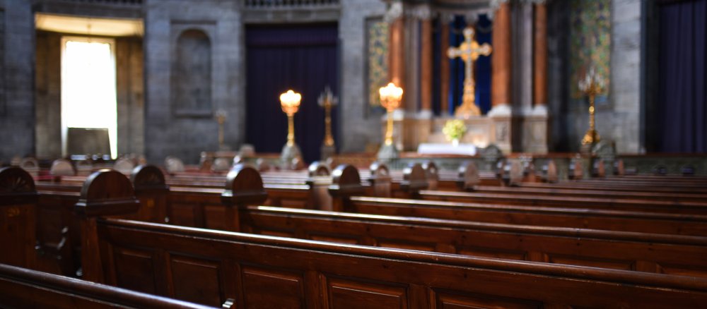 Der Blick über leere Bankreihen einer Kirche auf ein Kreuz.