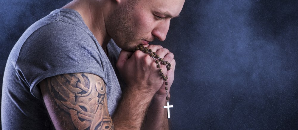 Ein Mann mit einem Tattoo auf dem Oberarm hat seine Hände zum Gebet gefaltet.