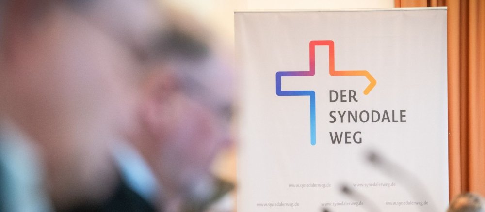 Stellwand mit dem Logo des Synodalen Wegs und Teilnehmer unscharf im Vordergrund