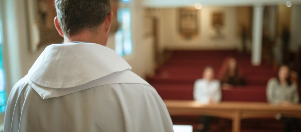 Priester steht vor einer Kirche mit drei Gottesdienstbesuchern