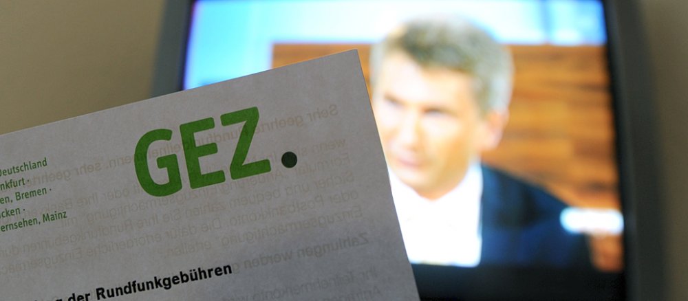 Eine Hand hält eine Rechnung der Gebühreneinzugszentrale der öffentlich rechtlichen Rundfunkanstalten (GEZ) für Radio und Fernsehen vor einen Fernseher.
