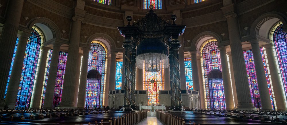 Bunte Glasfenster hinter dem Hauptaltar der Basilika Notre-Dame-de-la-Paix. Die Kirchenbänke davor sind leer.