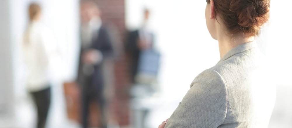 Eine Frau im Business-Outfit beobachtet ein Gespräch unter Kollegen.
