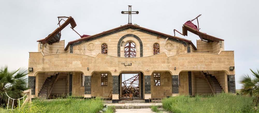 Außenanlage einer teilweise zerstörten christlichen Kirche in Karakosch, Irak. Die Fassade ist stark beschädigt, im Hintergrund liegen Trümmer.
