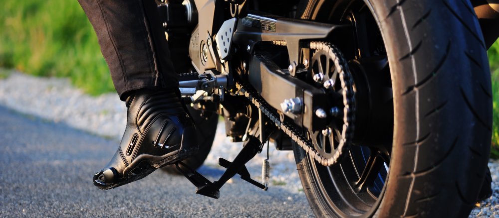Der Hinterreifen eines Motorrads steht auf dem Asphalt. Am linken Bildrand sieht man noch den Fuß des Fahrers in seinem schweren Stiefel.