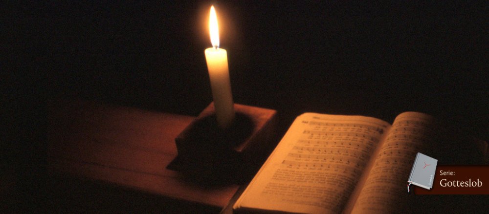 Ein Gesangbuch liegt aufgeschlagen auf einer Kirchenbank und wird nur vom Schein der kleinen Kerze links daneben beleuchtet.