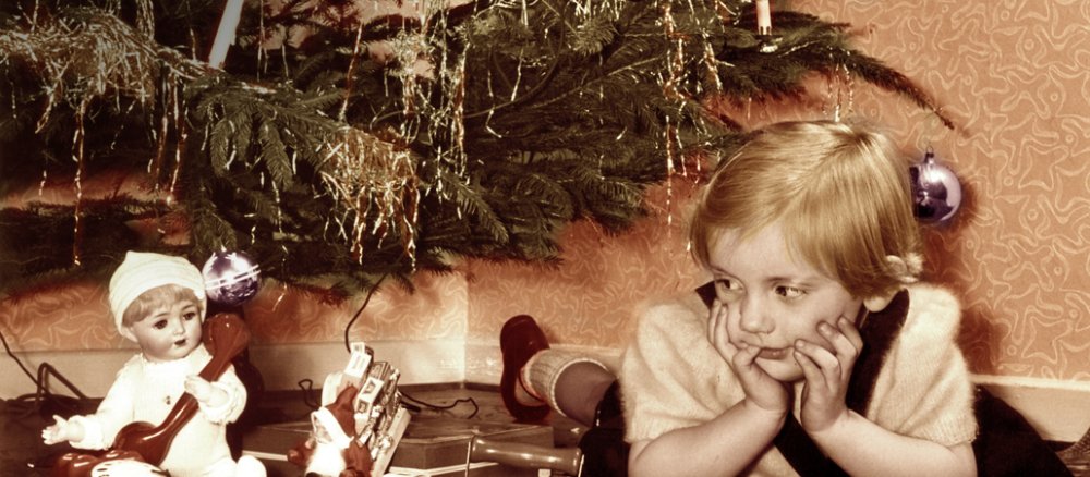 Ein kleines Mädchen liegt am Heiligen Abend 1955 unter dem Weihnachtsbaum und schaut auf die Geschenke, darunter eine Puppe, ein Kindertelefon und eine kleine Spielzeugeisenbahn.