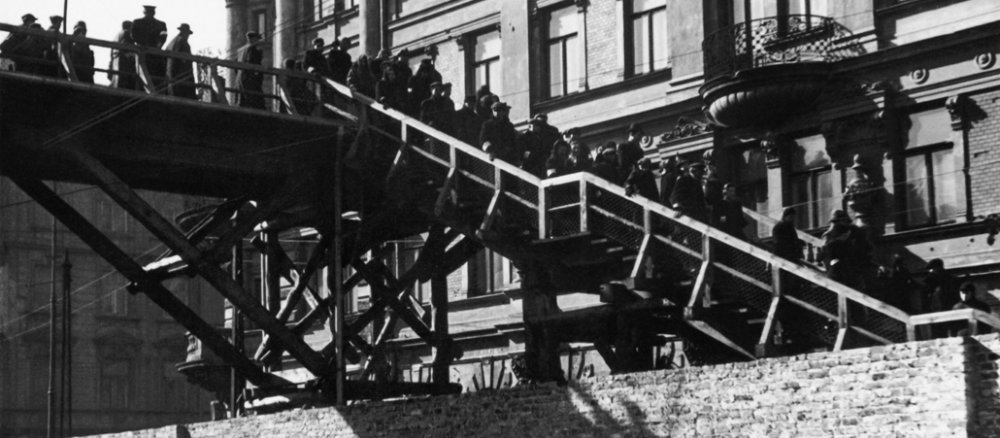 Mehrere dunkel gekleidete Menschen (Juden) gehen über eine Brücke, die am rechten Bildrand hinter einer Mauer verschwindet.