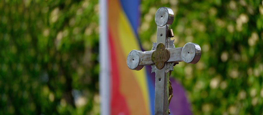 Ein Kreuz steht bei einem Open-Air-Segnungsgottesdienst für Liebende vor der Kirche Christi Auferstehung. Im Hintergrund ist eine Regenbogenfahne zu sehen.