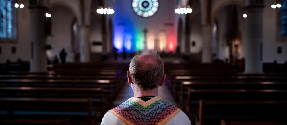 Pfarrer Heinrich Plaßmann steht in der Kirche und trägt eine Stola in den Farben des Regenbogens