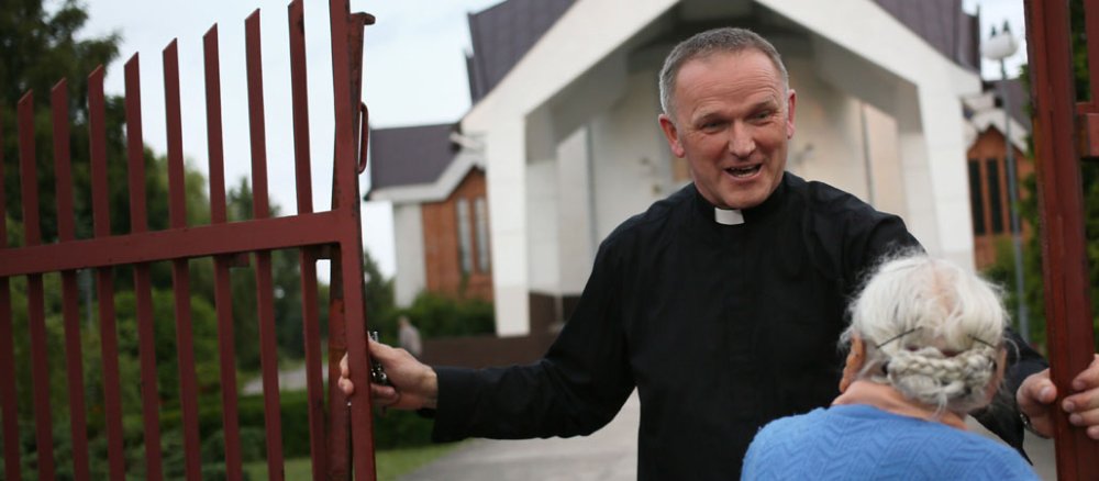 Am Eingangstor zu einem Kirchhof spricht ein Priester mit einer alten Frau