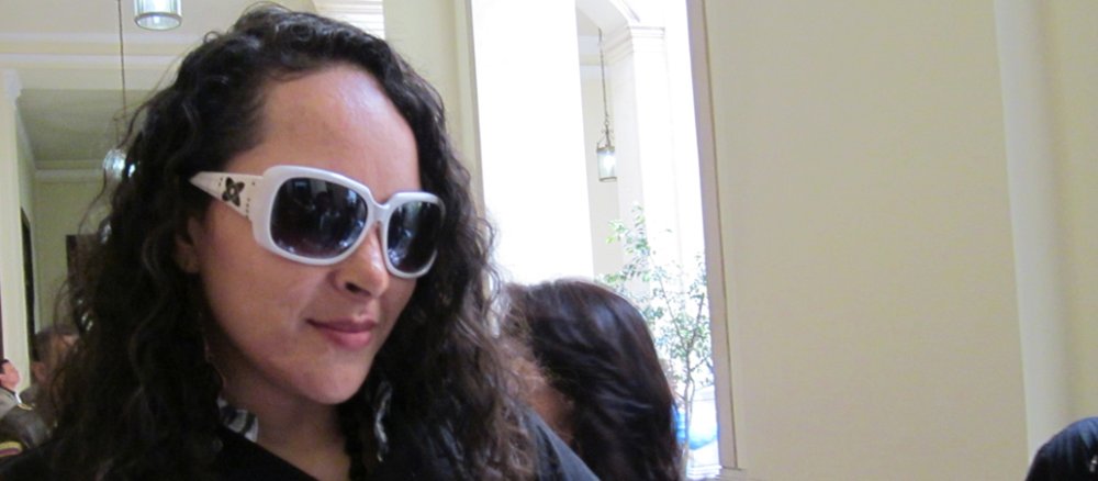 Eine Frau steht in einem beschrifteten T-Shirt und eine Sonnenbrille, die große Teile des Gesichts bedeckt, im lichtdurchfluteten Flur - vermutlich eines Gerichts- oder Verwaltungsgebäudes.