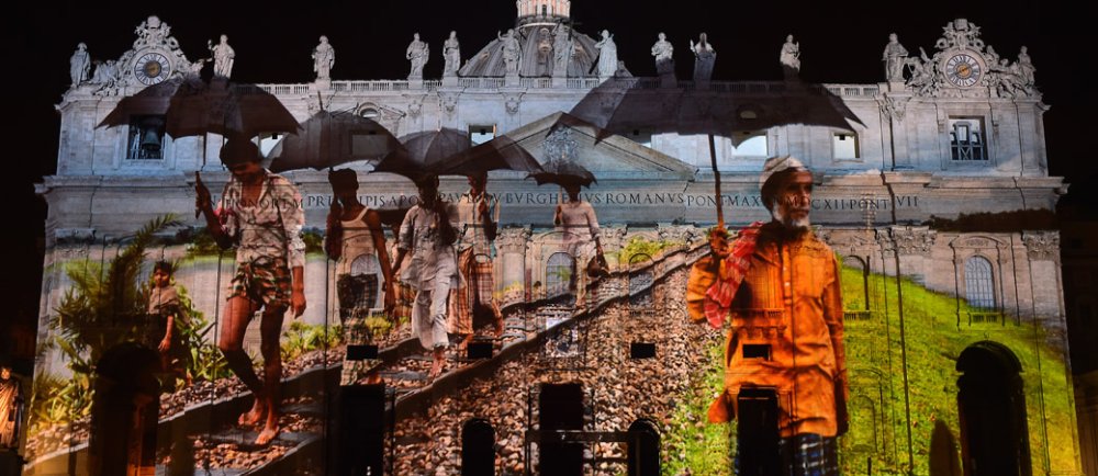 Eine Lichtinstallation zeigt Menschen auf der Fassade des Petersdoms.