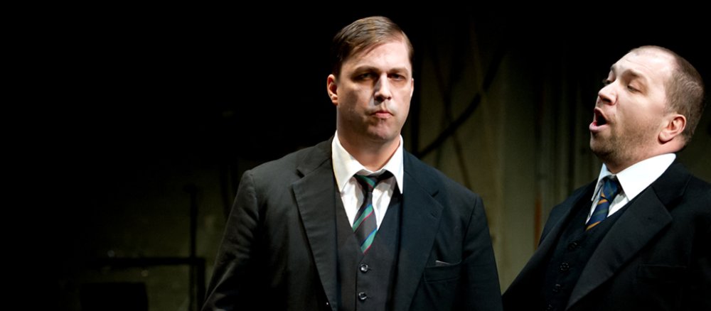 Zwei Schauspieler stehen in schwarzen Anzügen auf der Bühne, einzig die Gesichter heben sich vom dunklen Hintergrund ab. Der linke von beiden spielt Dietrich Bonhoeffer.