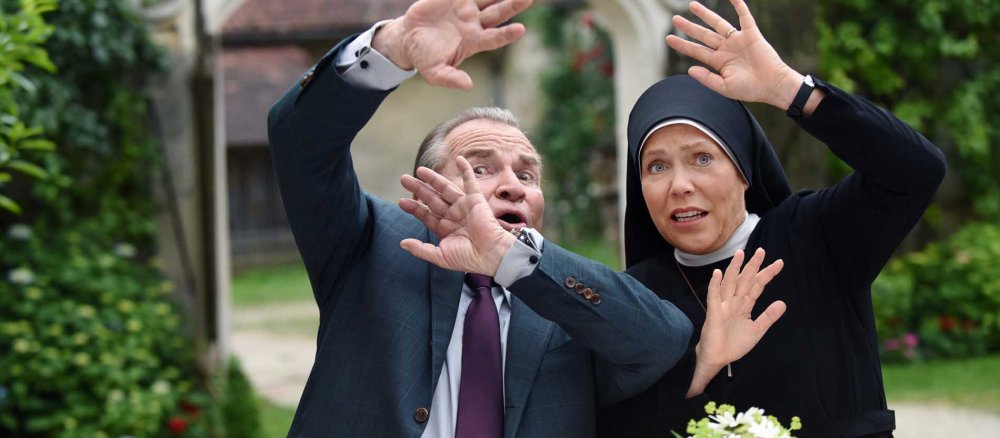 Ein Mann und eine Nonne stehen an einem Tisch und heben abwehrend die Arme.