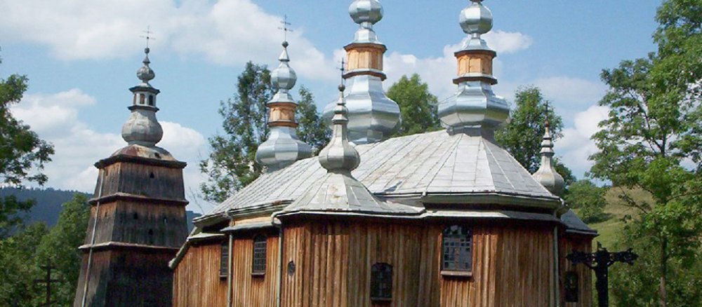 Eine Holzkirche im mit mehreren Türmen vor blauem Himmel auf einer grünen Wiese.