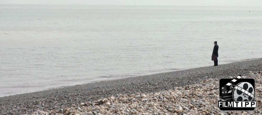 Mr. May steht am Wasser und schaut in die Ferne.