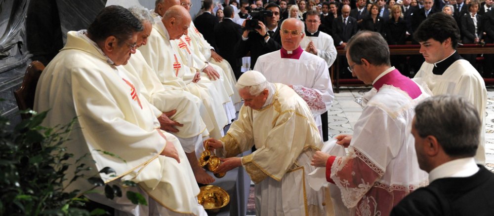 Fünf Karindäle sitzen in weißen Gewändern auf einer Bühne. Vor ihnen steht Papst Benedikt XVI. und füllt Wasser in einen goldenen Behälter um mit der Rituellen Fußwaschung an Gründonnerstag zu beginnen.