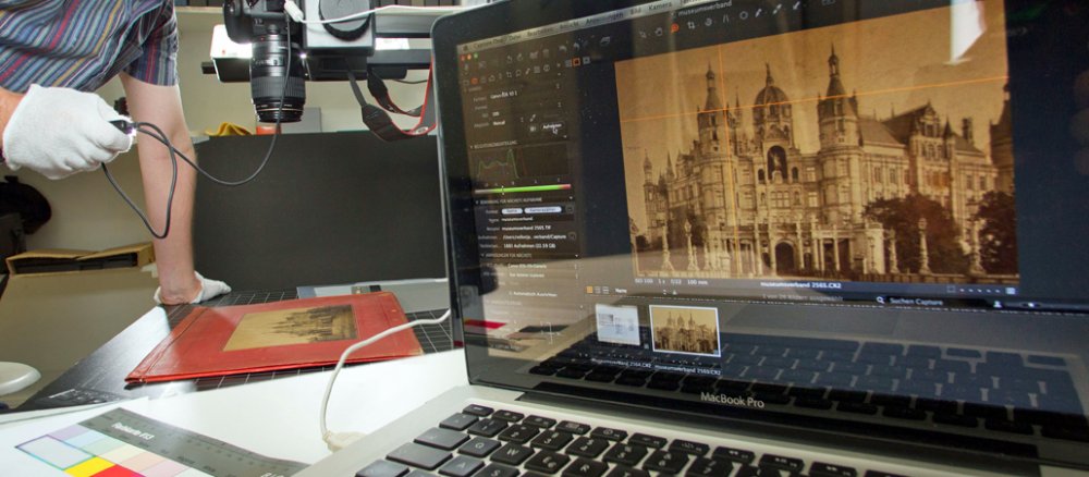 Links fotografiert eine kopfüber auf dem Stativ hängende Kamera ein Bild, das auf dem Tisch liegt. Rechts auf dem PC-Bildschirm ist es groß zu sehen: es ist das Schweriner Schloss.