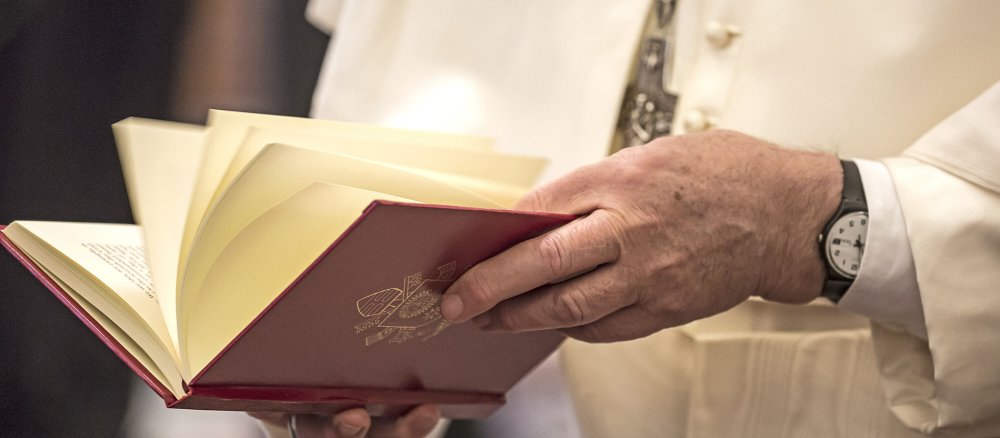 Eine Hand im päpstlichen Gewand blättert in einem roten Buch