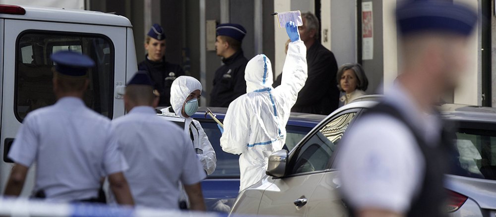 Im Vordergrund steht unscharf ein Polizisit, im Hintergrund stehen Ärzte in Schutzanzügen vor einem Krankenwagen.