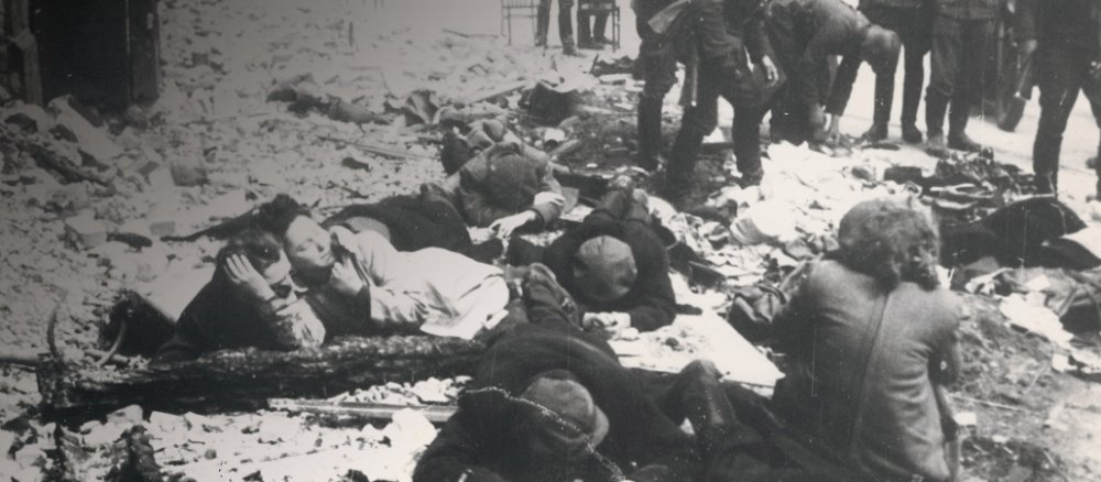 SS-Soldaten bewachen jüdische Widerstandskämpfer die sich im Schutt eines zerstörten Hauses auf den Boden ducken.