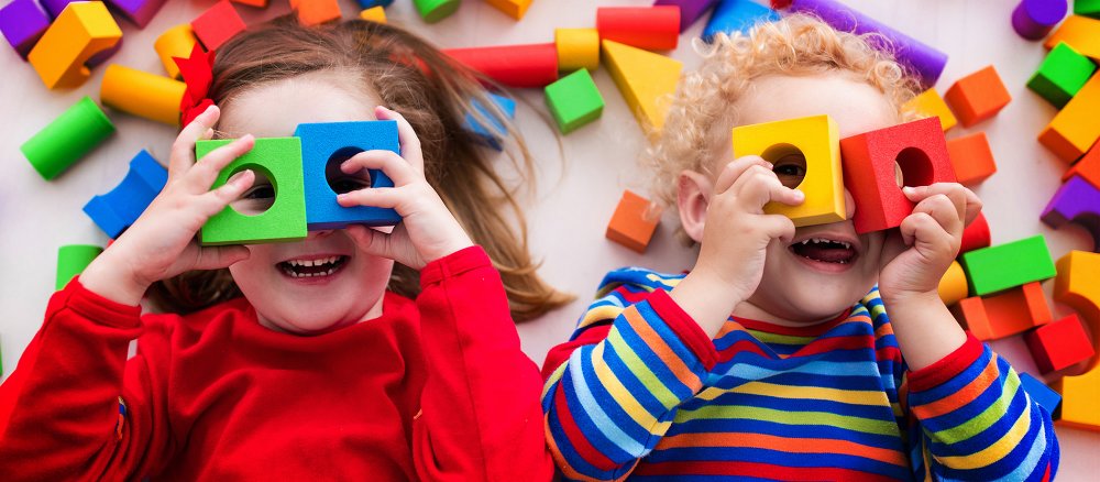 Zwei Kinder liegen rücklings auf dem Boden und spielen mit bunten Bauklötzen. Beide schauen durch je zwei Klötze mit Loch, die sie sich wie eine Brille vor die Augen gelegt haben.
