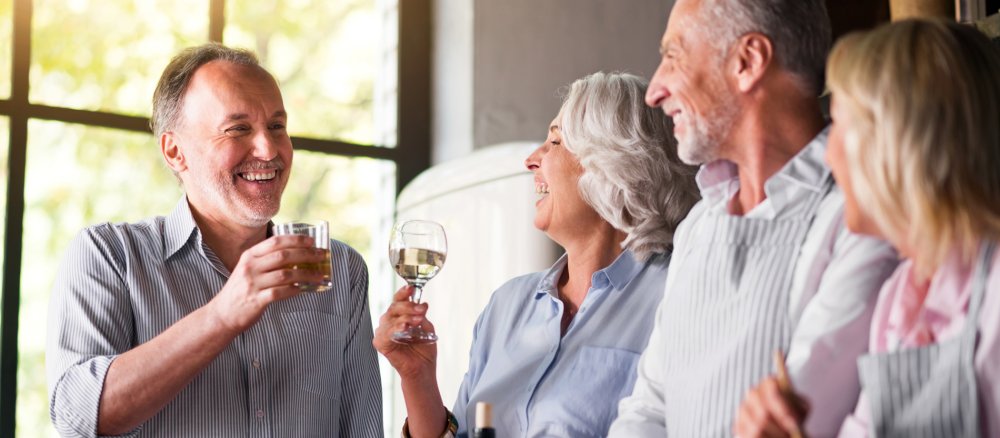 Zwei ältere Paare (m/w) stoßen in einer Küche mit Weißwein an.