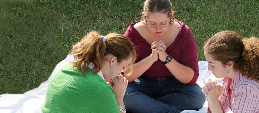 Drei junge Frauen sitzen auf einer Picknickdecke im Gras und haben die Hände gefaltet und die Augen geschlossen.