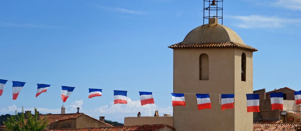 KIrchturm, Fahnenkette der Französichen Fahne von links nachs rechts durch das Bild, vor blauem Himmel, Häuserdächer