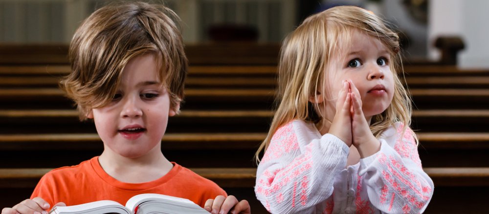 Zwei kleine Kinder sitzen in der Kirchenbank. Der Junge links schaut runter in ein Gotteslob, das Mädchen rechts hat die Hände gefaltet und blickt zum Altarraum.