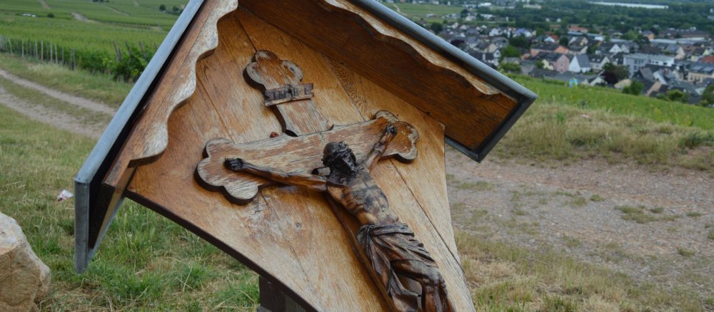 Wegkreuz mit Jesusfigur aus Holz am Wegesrand mit Blick auf ein Weindorf im Hintergrund.