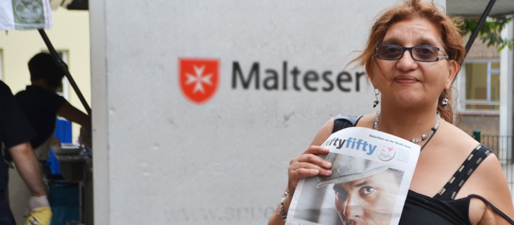 Füllige Frau mit Trägertop, Brille und Schmuck hält Obdachlosenzeitung in die Kamera