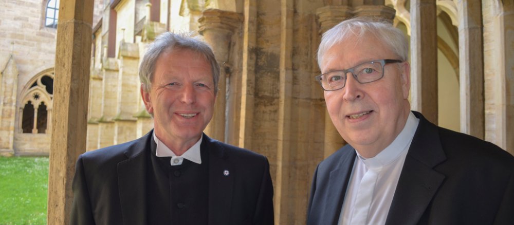 Zwei weißhaarige Männer stehen in dunklen Anzügen im Säulengang einer Kirche.