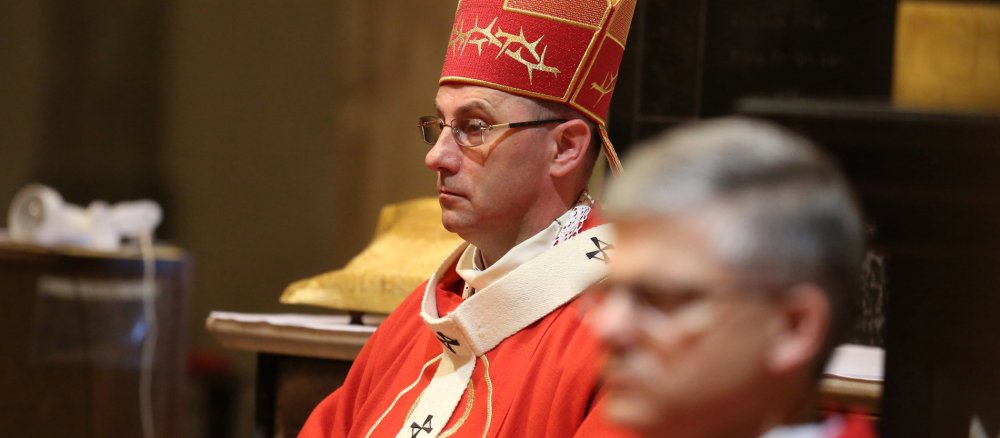 Ein gut 50-jähriger, schlanker Mann, sitzt in einem roten Gewand und mit Mitra auf dem Kopf in einer Kirche.