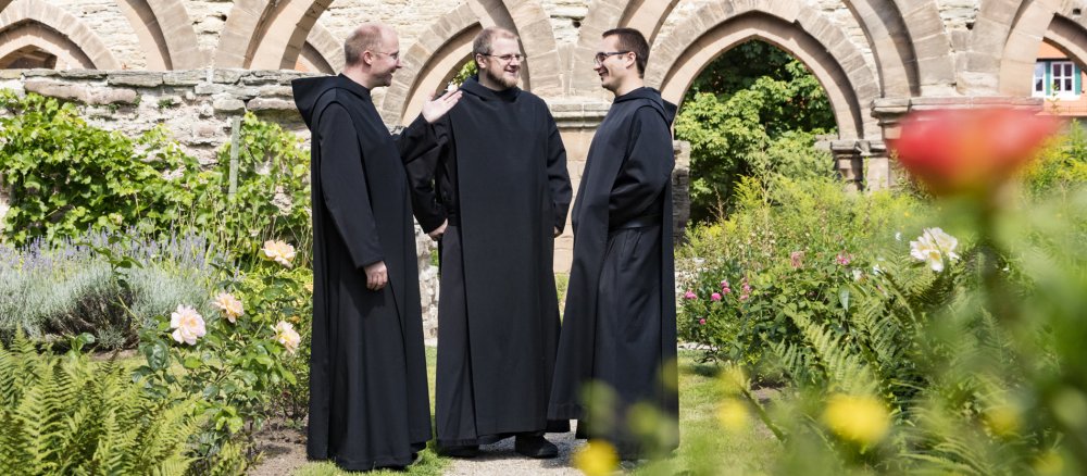 Drei Benediktiner-Mönche im schwarzen Habit stehen in einem Klostergarten zusammen.