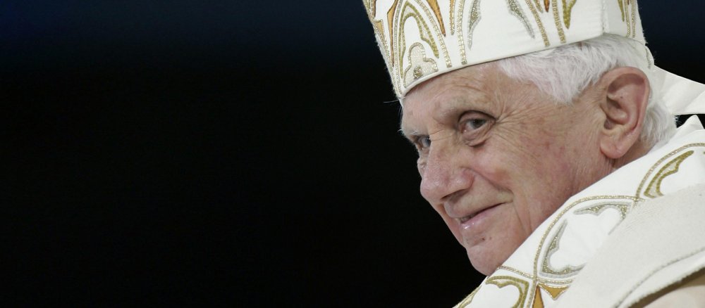 schwarzer Hintergrund, am linken Bildrand das Gesicht des lächelnden Benedikt XVI.