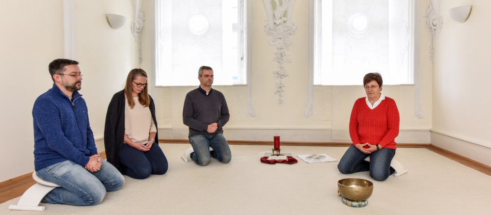 Vier Menschen knien meditierend im Halbkreis in einem lichtdurchfluteten Raum.