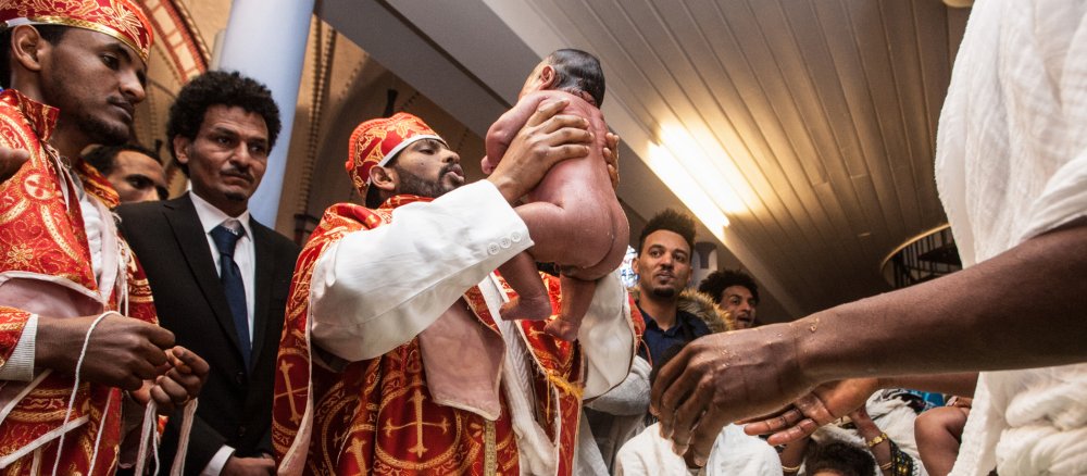 Ein Priester hält einen nassen, nackten Säugling in die Höhe