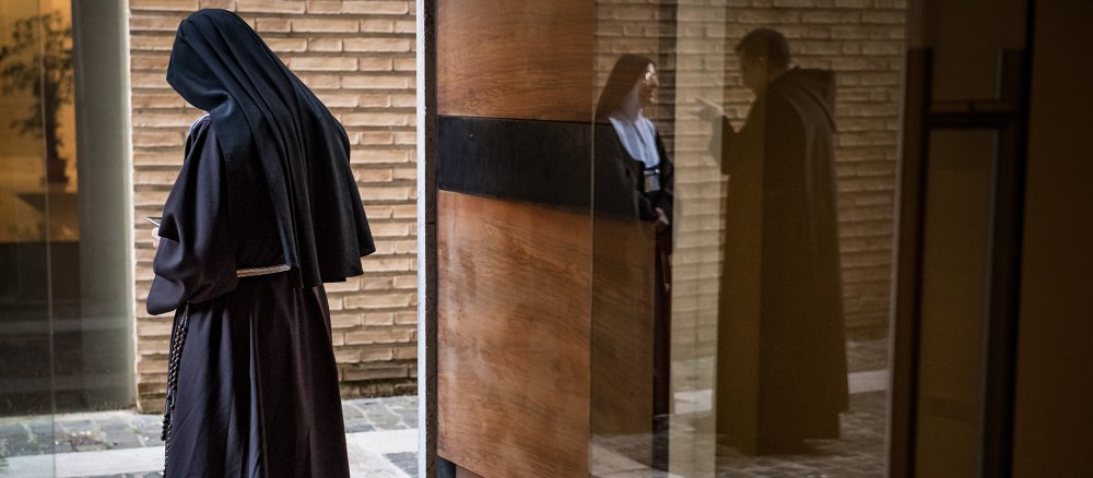 Eine Ordensfrau mit schwarzem Habit und Schleier steht mit gesenktem Kopf. Im Fenster spiegeln sich eine Ordensschwester und ein Ordensmann im Gespräch.