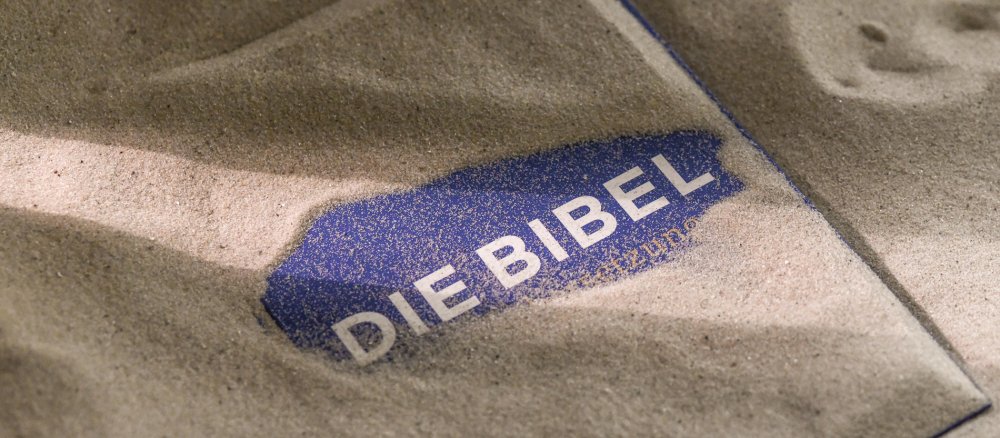 Eine Bibel ist mit Sand bedeckt, sodass nur der Bibel-Schriftzug zu sehen ist